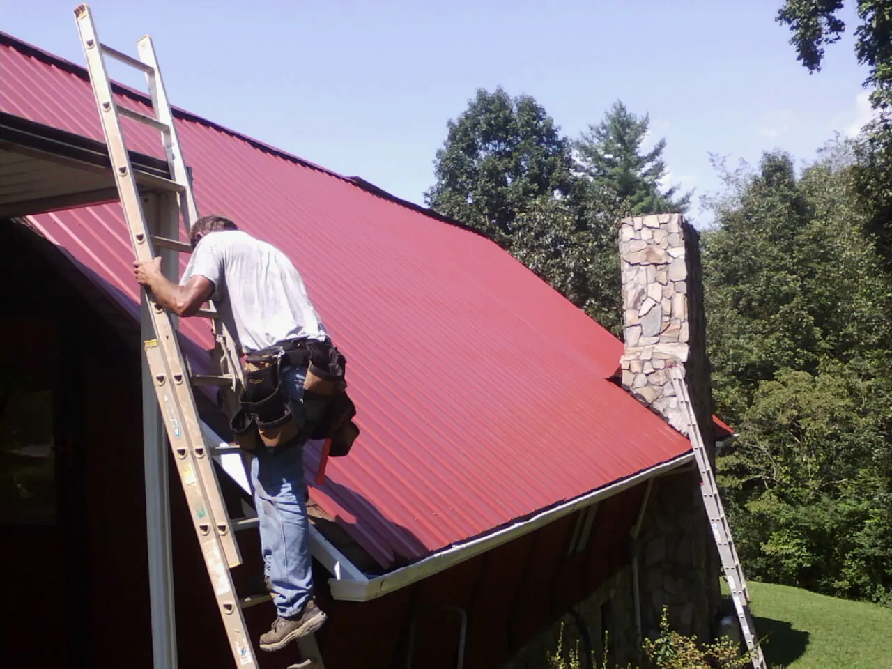 Roofer on a ladder beside a red metal roof and stone chimney — Soffit & Fascia Repair by Roofing Brothers in Clifford